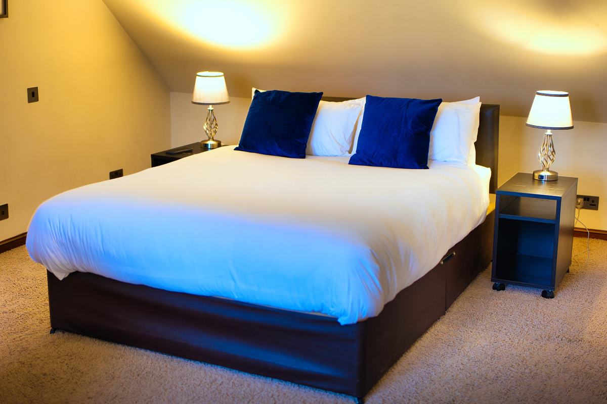The Royal Clifton Suite: a king-size bed with crisp white linen and navy velvet cushions under a sloping ceiling, lit by two bedside lamps